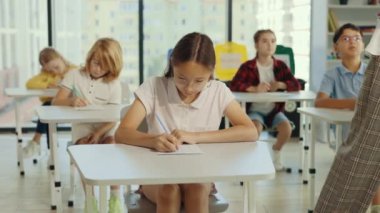 Beautiful brunette asian school girl wearing glasses sits at a first desk in the classroom and fills out tests thinks about answers, and the teacher going through on the background