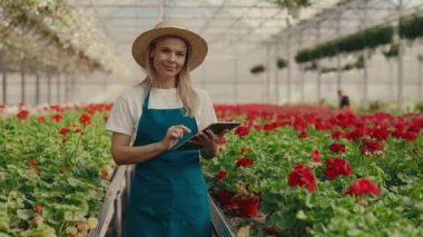Female gardener wearing hat holding tablet and calculate something standing near the shelf with flowers and plants looking at the camera. People and professions concept