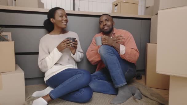 幸せなアフリカ系アメリカ人のカップルの床に熱いお茶やコーヒーを飲み 彼らの新しい家について話して座っている 新しい住宅移転と不動産コンセプト — ストック動画