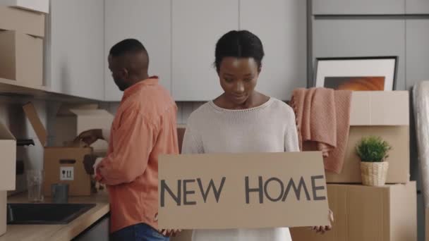 カメラを見て笑顔の新しいキッチンに立って段ボール箱の新しい家を保持陽気なアフリカ系アメリカ人女性の肖像画 人と移転の概念 — ストック動画