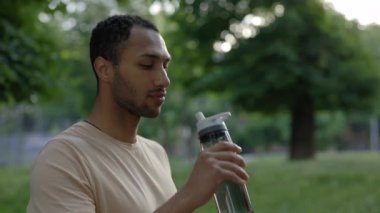Afro-amerikan adam spor şişesinden su içiyor. Erkek koşucu koşudan sonra şehir parkında duruyor. Spor ve sağlıklı yaşam tarzı kavramı