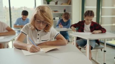 Kırmızı liseli gözlüklü çocuk masaya oturmuş dikte yazıyor. Okula geri dön. Halk ve eğitim kavramı