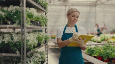 Çiçeklerle dolu arabanın yanında duran bahçıvan bitkiler hakkında bir şeyler yazdı. Kadın sera işçisi, mutlu botanikçi yakın plan, küçük işletme sahibi. Halk ve meslek kavramı