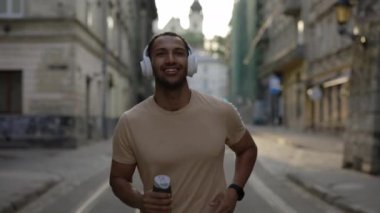 Afrika kökenli Amerikalı bir adam eski kasaba sokaklarında koşuyor kulaklık takıyor ve müzik dinliyor. Mutlu erkek sporcu yaz sabahı antrenmanının tadını çıkarıyor. Spor ve sağlıklı yaşam tarzı kavramı