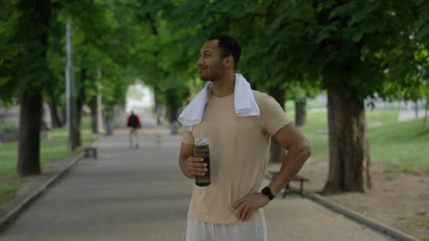 スマートウォッチを装着したスポーツボトルを持ったカメラを見ているアフロアメリカ人男性の肖像画は 訓練後 公園で彼の首の周りにタオルを持っています スポーツと健康的なライフスタイルのコンセプト — ストック動画