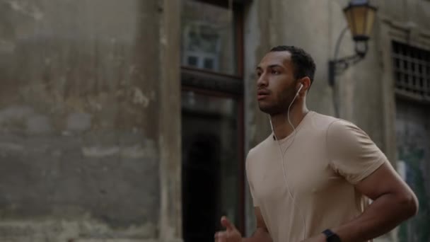 非洲裔美国人穿着耳机在老城区街道上跑步 听音乐的侧视图 体育与健康生活方式概念 — 图库视频影像