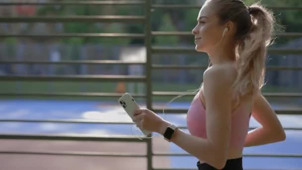イヤフォンとスマートウォッチを身に着けて実行している若い美しい女性の側のビューでは 彼女の手の中に携帯電話を保持カロリーを計算します スポーツと健康的なライフスタイルのコンセプト — ストック動画