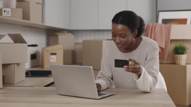 アフリカ系アメリカ人の女性は 新しい家でカードとラップトップを使用してインターネット経由で新しいアパートから購入するために支払います 女性はクレジットカードで支払っている 移転だ 人と技術の概念 — ストック動画