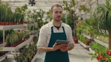 Elinde dijital tabletle kameraya bakıp gülümseyen yeşil bahçe çalışanı portresi. Erkek bahçıvan, bitki ve çiçek serasında çalışıyor. Halk ve meslek kavramı