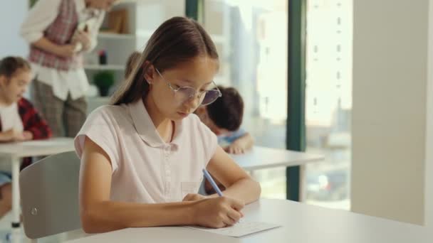 小学校での試験 ガラスを身に着けている美しいブルネットのアジアの女子高生は教室の最初の机に座ってテストを記入します 教育子供 — ストック動画