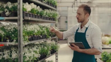 Erkek bahçıvan çiçeklerle dolu el arabasının yanında duruyor ve kalitesini kontrol ediyor ve dijital tablete yazıyor. Halk ve meslek kavramı