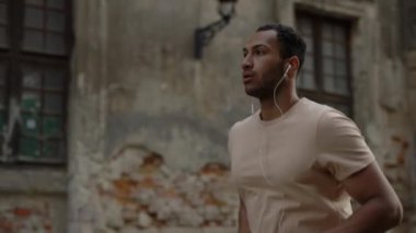 Eski kasaba sokaklarında kulaklıkla koşan Afro-Amerikan adamın yan görüntüsü. Koşucu koşarken kulaklıkla müzik dinliyor. Spor ve sağlıklı yaşam tarzı kavramı