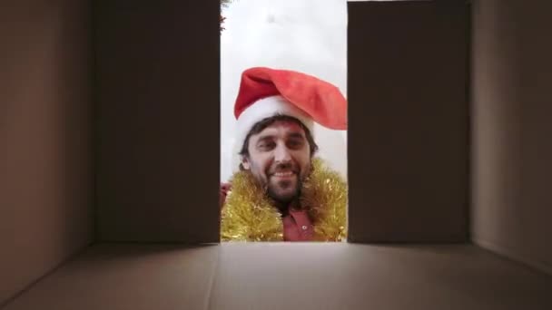 サンタの帽子に身を包んだ白人男性は 驚くべきクリスマスプレゼントを取るために段ボール箱の中に手を入れました 現在から男性の顔に喜びと満足 — ストック動画