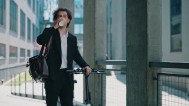 İş adamı iş merkezine resmi siyah takım elbiseyle gidiyor, elinde elektrikli bisikletle kahve içiyor. İş yerinin yakınındaki erkek patron etrafa bakıyor. İnsanlar ve teknoloji konsepti.