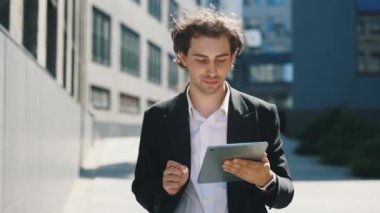 Dijital tablet kullanan yakışıklı beyaz adam, iş merkezinin yakınında yürüyor, mesajlaşıyor, uygulamaları kaydırıyor. İnsanlar ve teknoloji kavramı