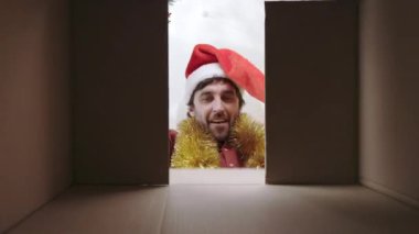 Samimi mutluluğa sahip neşeli sakallı adam karton kutunun içindeki Noel hediyesine tepki veriyor. Beyaz erkek, Noel Baba şapkalı. Evde karton paketi açıyor..