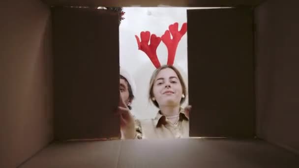 赤いサンタの帽子をかぶった正の白人カップルは クリスマスプレゼントを入れた段ボール箱を内側に展開しています 二人の陽気な家族の低角度ビュー — ストック動画