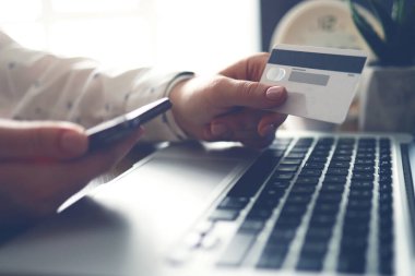 Female hands holding credit card and phone near the laptop. Close-up. Online payments. Banking transfer.