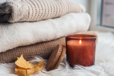 Burning candle on the windowsill, against the background of warm sweaters and autumn leaves.
