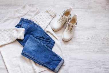 Womens autumn flat clothes with warm white sweater, blue jeans and white shoes, feminine fashionable outfit on white sheet, copy space