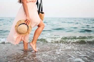 Woman in straw hat and pink dress on a tropical beach with brown bag.