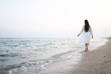 Güzel genç kadın uzun saçlı ve şapka uzun beyaz elbiseli plaj boyunca gider