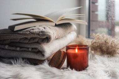Burning candle and sweaters with a book on the windowsill. Autumn mood.