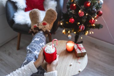 Girl holding a red cup with cocoa and marshmallows in her hands, cozy Christmas morning.
