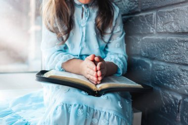 Little girl hands folded in prayer on a Holy Bible in church for faith concept in vintage color tone.