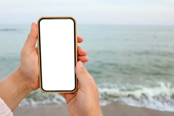 Hand showing a blank smart phone on the beach with the sea in the background. White screen mock up