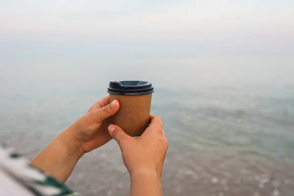 Coffee at the sea. Young woman holds a disposable cup of coffee on on a sunny morning at the beach.