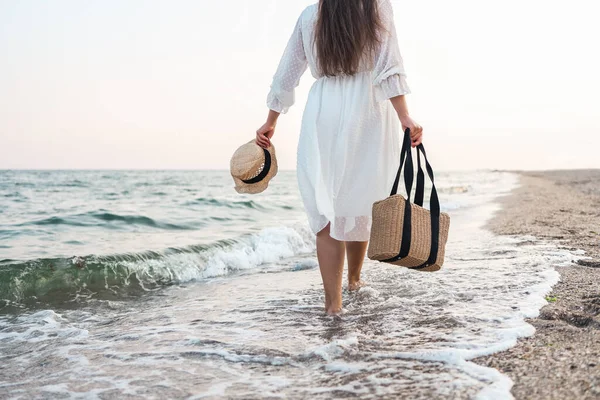 Woman in straw hat and white dress on a tropical beach with brown bag.