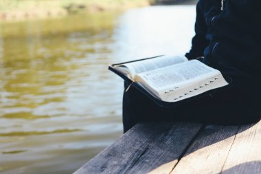 Child reads Bible book while sitting near pond. Christian illustration. Place for text.