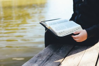 Child reads Bible book while sitting near pond. Christian illustration. Place for text.