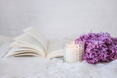 An open book with a lilac branch and a candle on a white fluffy plaid. Beautiful card. Good morning concept.