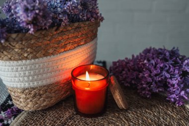 A burning candle with a wicker basket of lilacs. Beautiful aesthetic photo.