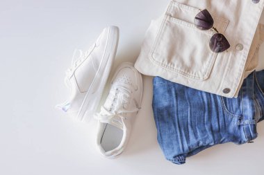 Stylish women's clothing flat lay. White sneakers, jeans and a jacket on a white background top view.