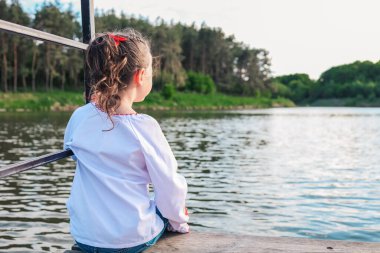 The child sits on a wooden bridge near the river, enjoys the beauty of nature.