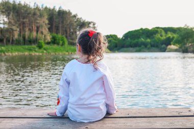 The child sits on a wooden bridge near the river, enjoys the beauty of nature.