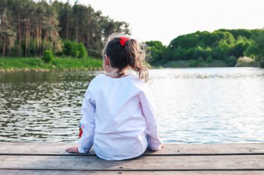 The child sits on a wooden bridge near the river, enjoys the beauty of nature.