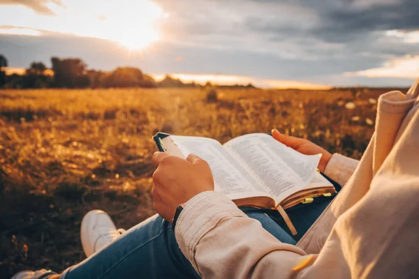 Christian woman holds bible in her hands. Reading the Holy Bible in a field during beautiful sunset. Concept for faith, spirituality and religion. Peace, hope