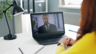 Modern ofisteki webcam konferansı sırasında sakallı bir meslektaşla konuşan olgun bir kadının omuzlarının üzerinden. Beyaz masadaki dijital dizüstü bilgisayara odaklan.
