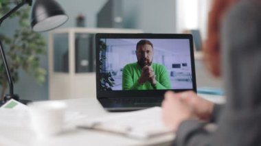 Modern dizüstü bilgisayardaki görüntülü sohbet sırasında masa başında oturan ve erkek meslektaşımla konuşan kadın ofis müdürünü kapatın. Yeşil kazaklı sakallı adamla ekrana odaklan..