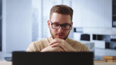 Ofis masasında oturup video konferansı sırasında konuşan gözlüklü yakışıklı bir genç. Kafkas erkek çalışanı meslektaşlarıyla çevrimiçi iletişim kurmak için kablosuz bilgisayar kullanıyor.