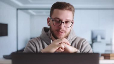 Modern dizüstü bilgisayardaki online toplantı sırasında ofiste oturan kulaklıklı yakışıklı genç adam. Odaklanmış erkek çalışanlar günlük giysiler içinde elleri ile hareket ederken çalışma stratejisini açıklıyorlar..
