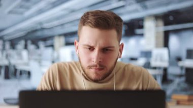 Modern kulaklıklı yakışıklı genç adam çalışma alanında otururken dizüstü bilgisayarında yaratıcı iş fikirleri yazıyor. Erkek çalışan yeni iş stratejisi üzerinde çalışıyor.