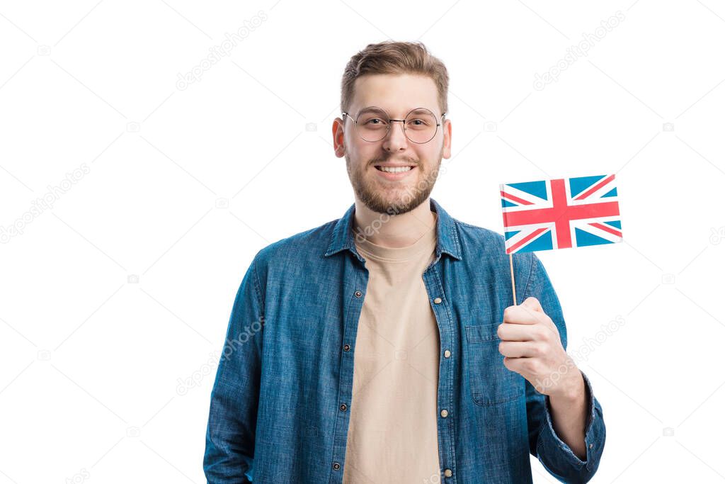 Patriotic young male holding national flag of UK while standing over white background. Studio portrait of proud englishman smiling sincerely. Patriotism and modern youth concept.
