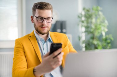 Caucasian male in formal wear sitting at workplace with modern smartphone and laptop. Positive young man browsing news while resting from work in office.