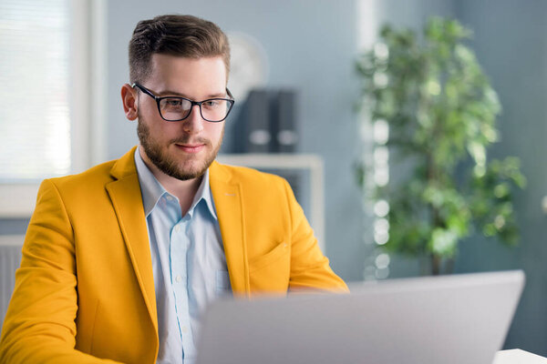 Positive bearded businessman sitting at office desk and working online. Handsome male typing creative business ideas on laptop in bright business center.