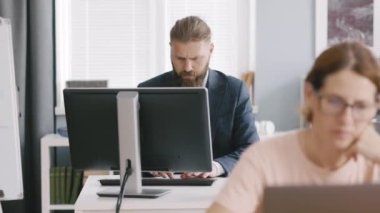 Bearded office manager in suit working on personal computer at modern office with other colleagues. Workplace, people and technology concept.